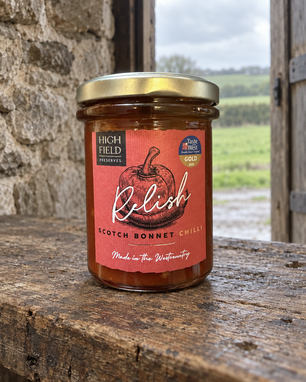 A jar of High Field Preserves Scotch Bonnet Chilli sauce sits on a rustic wooden table with a stone wall and an outdoor field in the background. The jar has a gold lid and a terracotta-colored label with a red pumpkin illustration and white script.