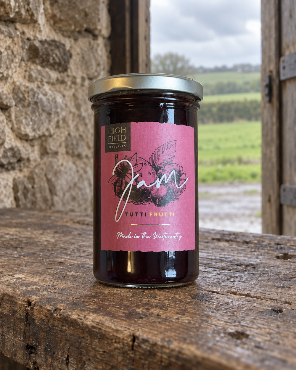 A glass jar of High Field Family Farm Jam, Tutti Frutti, sits on a rustic wooden table with a stone wall and an open doorway in the background.