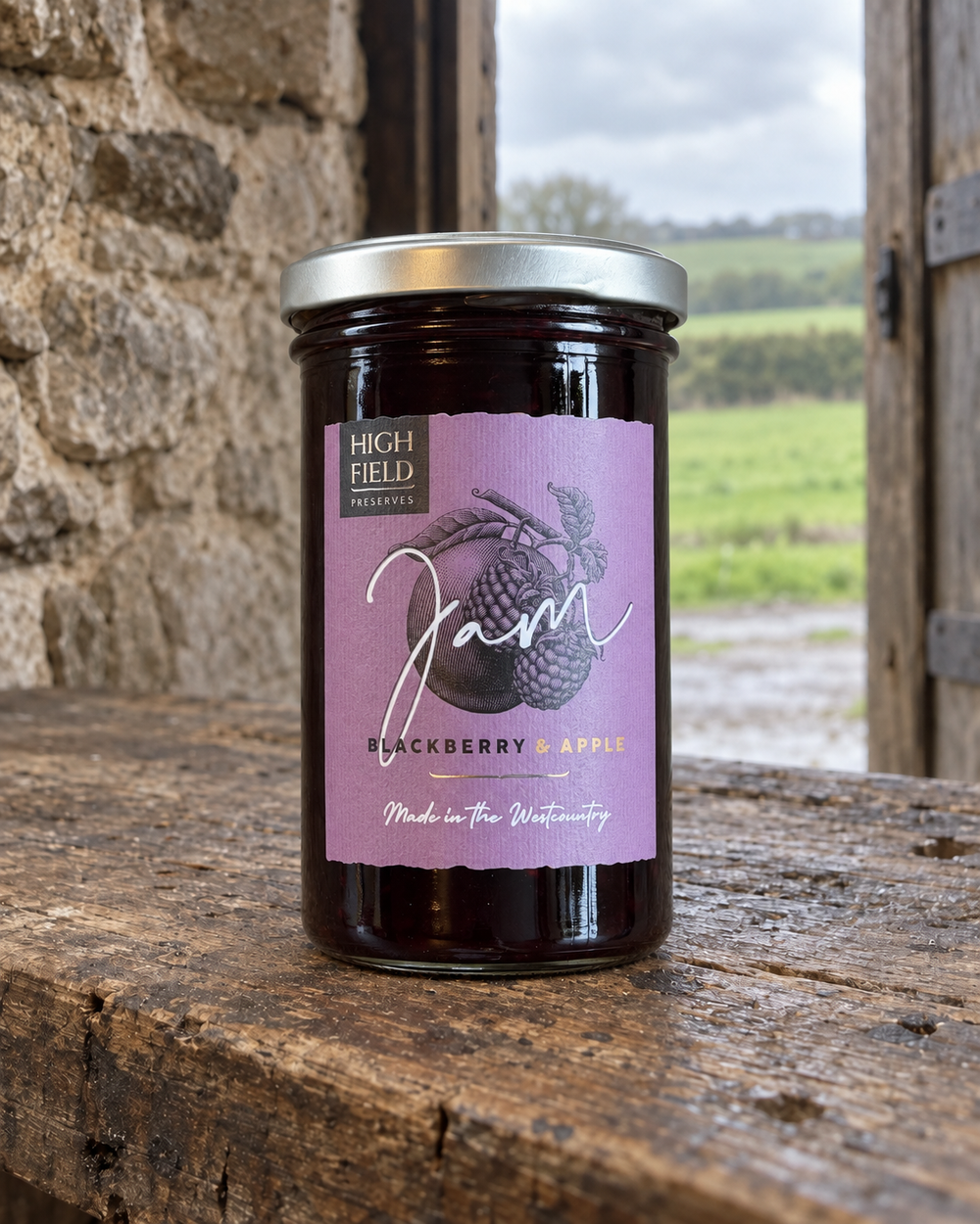 A glass jar of High Gold Millworks Blackberry & Apple jam sits on a rustic wooden surface in front of a stone wall, with an outdoor field visible in the background.
