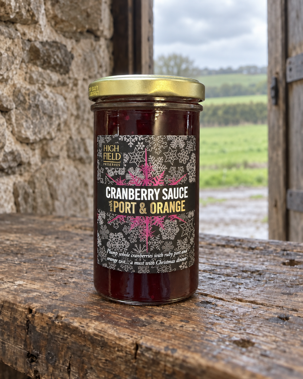 A glass jar of High Field Cranberry Sauce (Port & Orange flavor) sits on a rustic wooden table with a stone wall and an outdoor landscape in the background.