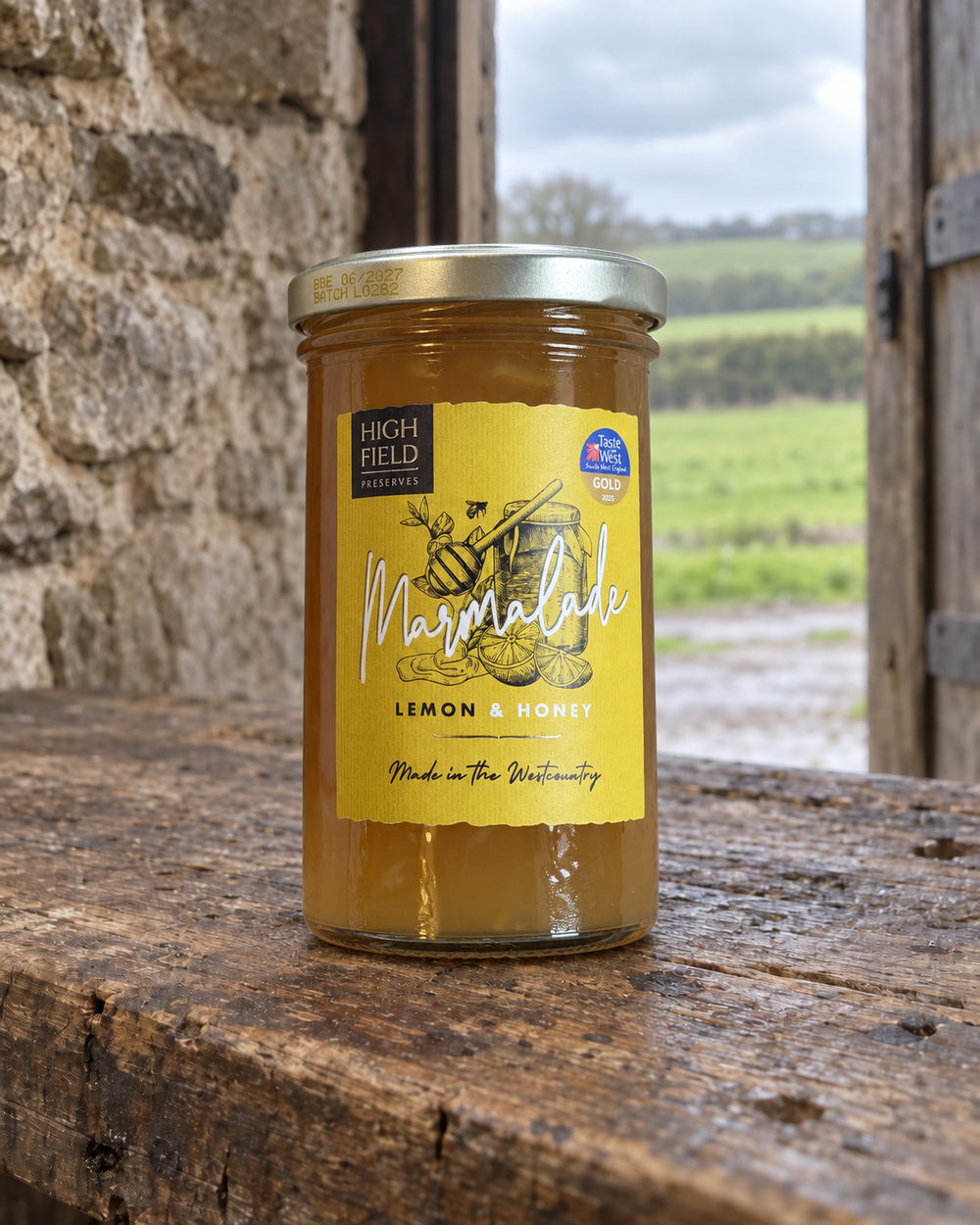 A jar of High Field Preserves  Marmalade (Lemon & Honey) sits on a rustic wooden table with a stone wall and a green field in the background.