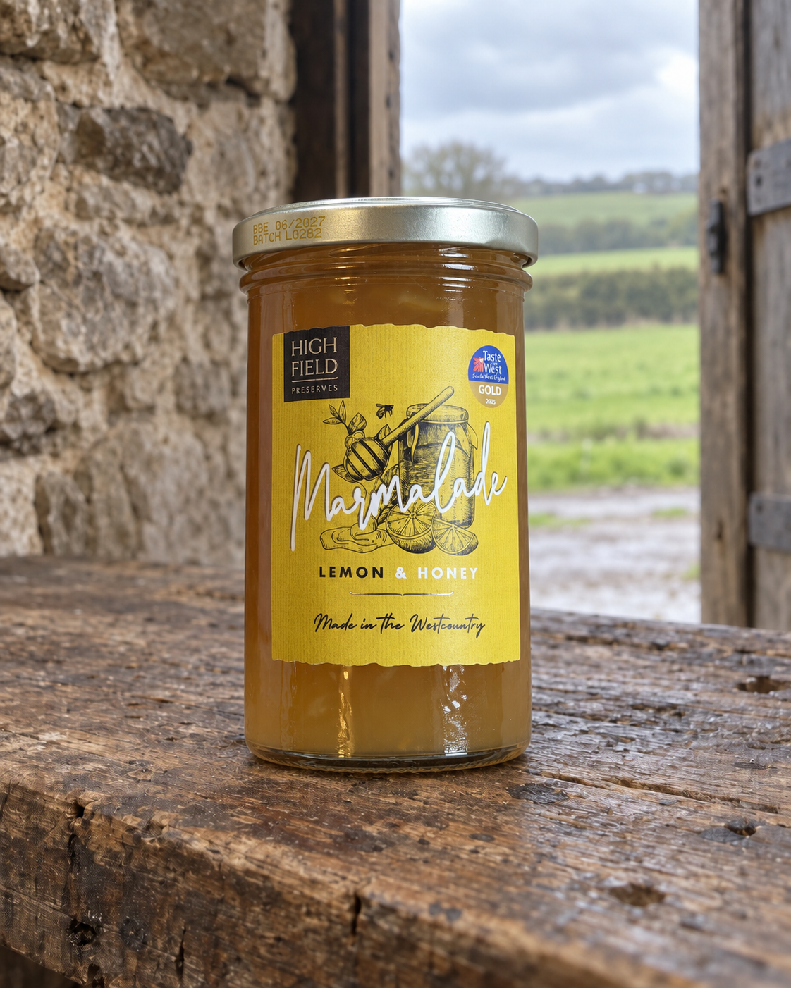 A jar of High Field Preserves  Marmalade (Lemon & Honey) sits on a rustic wooden table with a stone wall and a green field in the background.