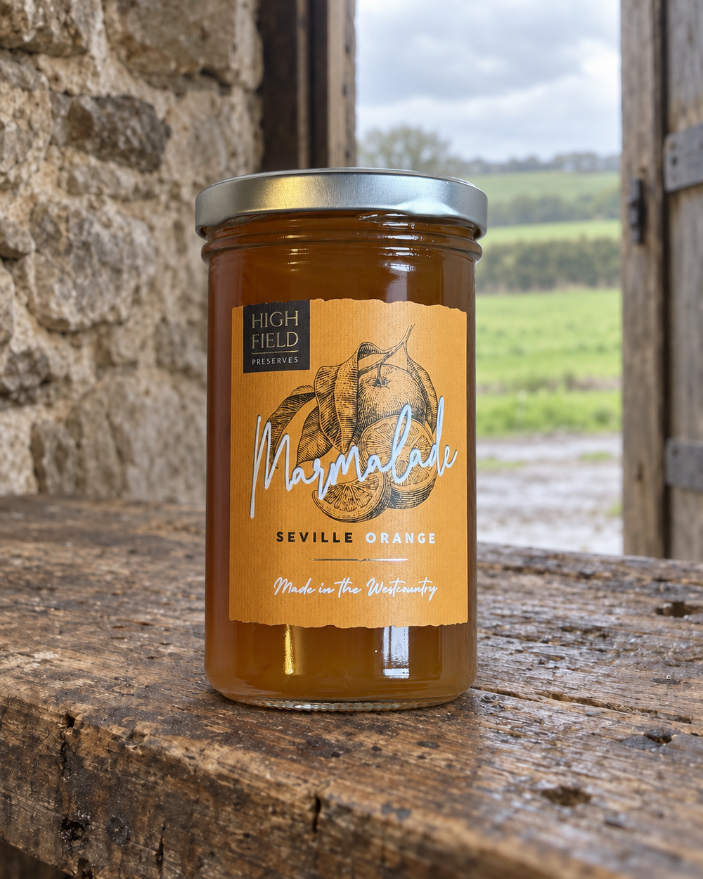 A glass jar of High Field Colchester Marmalade (Seville Orange) sits on a rustic wooden table with a stone wall and an open wooden door in the background, creating a natural, country-style setting.