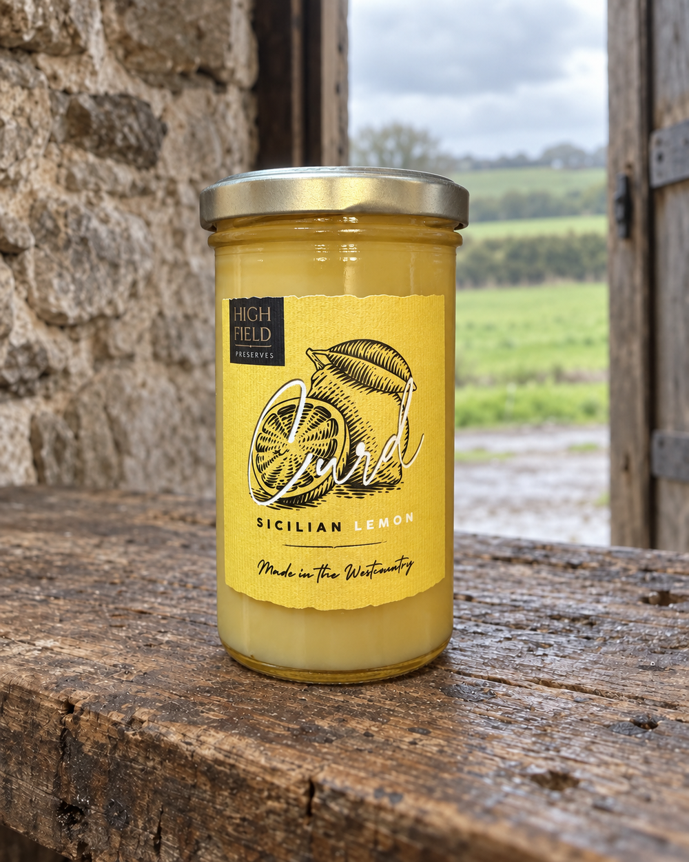 A glass jar of High Field Sicilian Lemon curd sits on a rustic wooden table with a stone wall and an outdoor field in the background. The candle wax is a pale yellow colour, and the label is a warm yellow with black text and an illustration of a lemon slice.