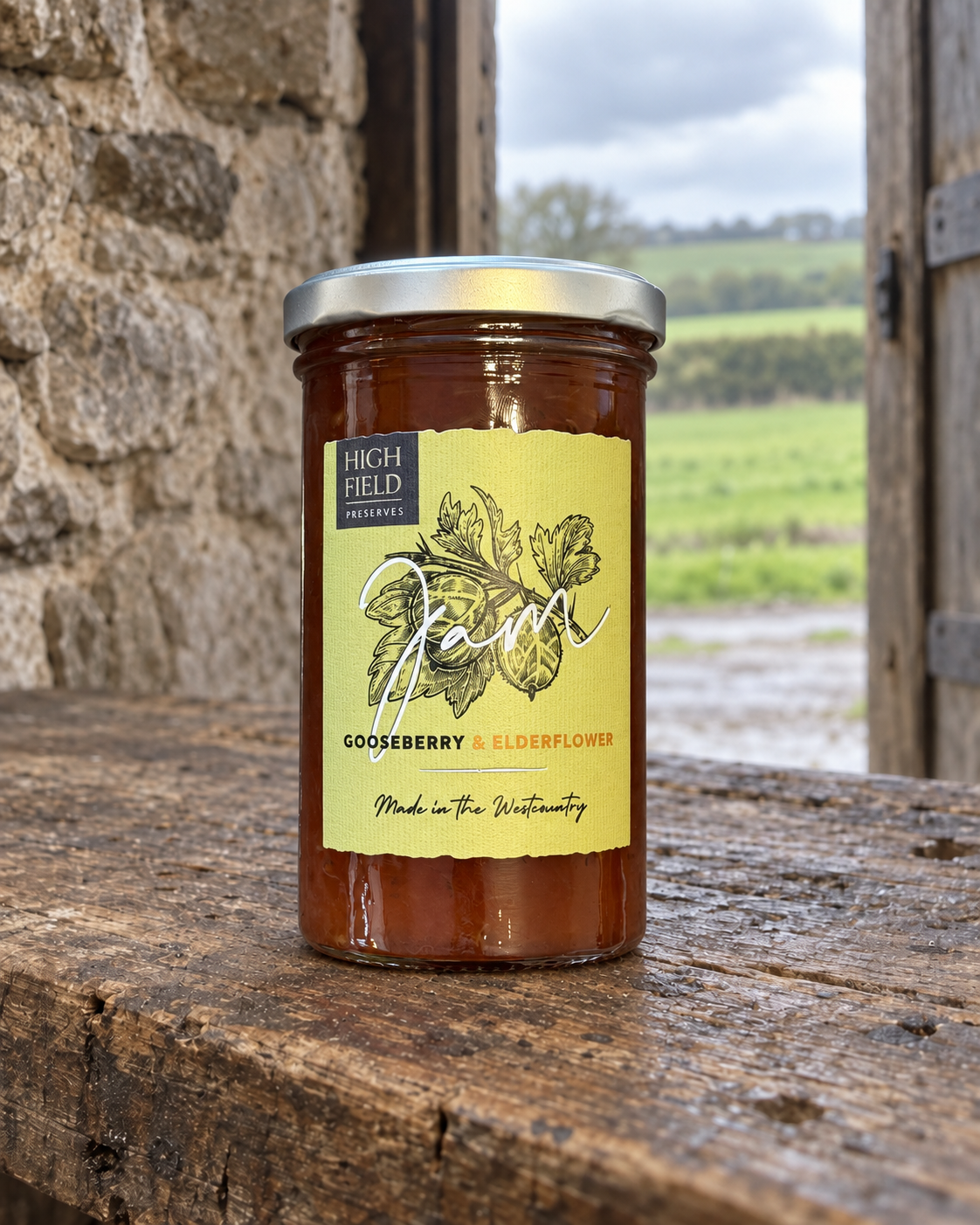 A jar of High Field Preserves Gooseberry & Elderflower jam sits on a rustic wooden table in front of a stone wall, with an outdoor field visible in the background.
